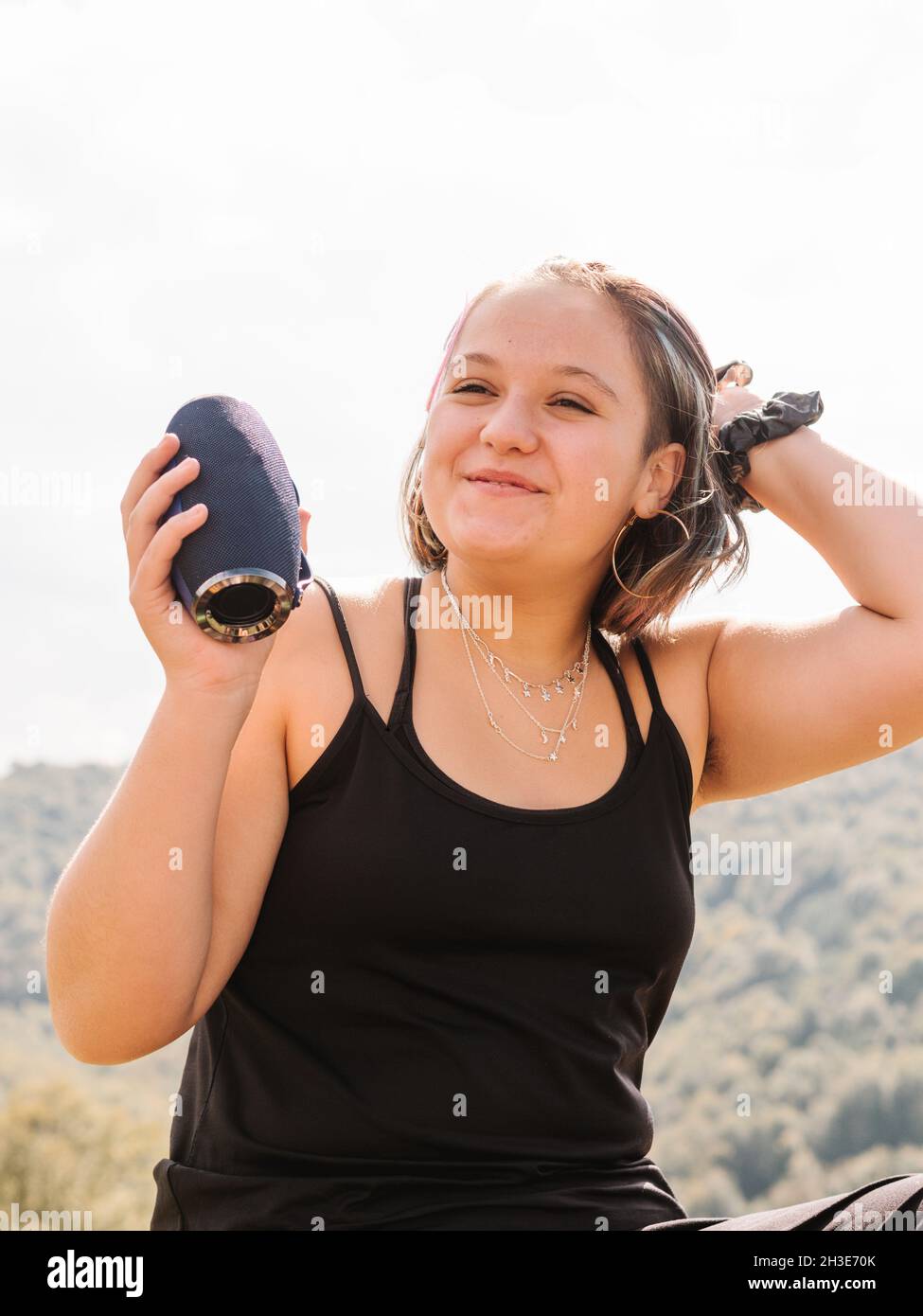 Positive Frau mit tragbarem bluetooth-Lautsprecher, die wegschaut, während sie auf bewaldeten Bergrücken mit grünen Bäumen in der Natur sitzt Stockfoto