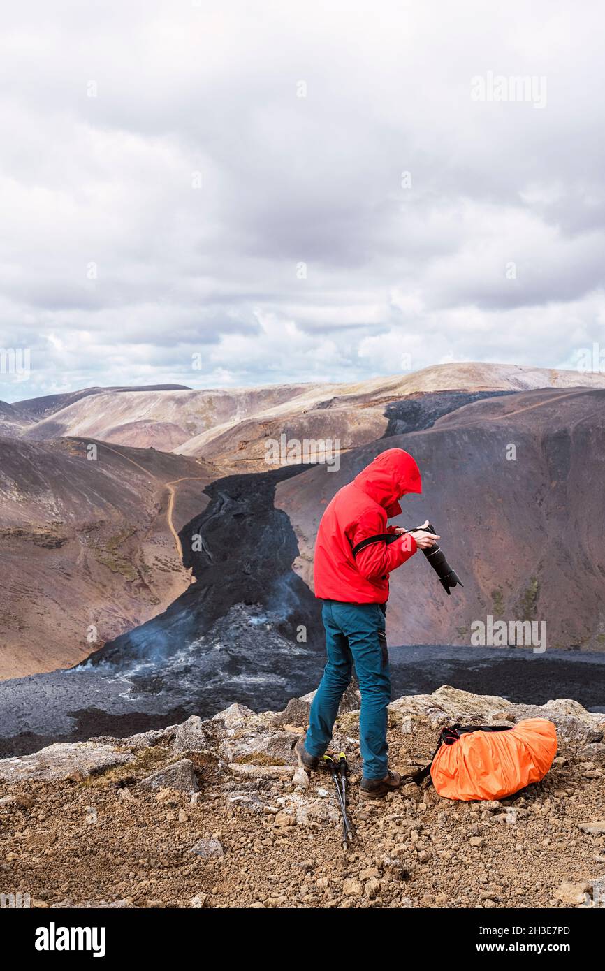 Seitenansicht eines männlichen Fotografen in Oberbekleidung, der auf einer felsigen Klippe in der Nähe des aktiven Vulkans Fagradalsfjall mit schwarzer Lava in Island tagsüber steht Stockfoto