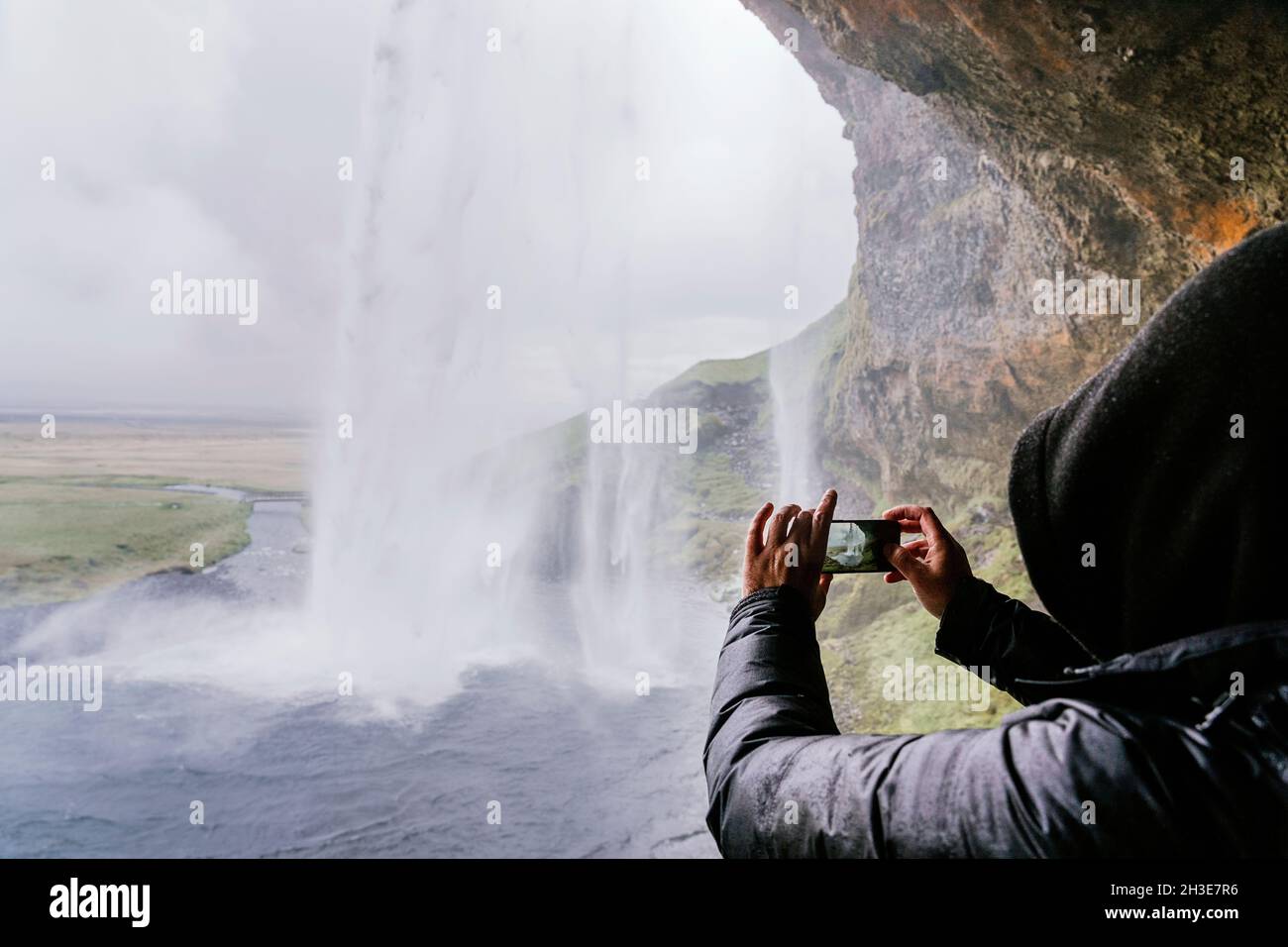 Seitenansicht eines nicht erkennbaren Reisenden in warmer Oberbekleidung und Hoodie, der während der Reise in IC den malerischen Seljalandsfoss-Wasserfall auf dem Smartphone fotografiert Stockfoto