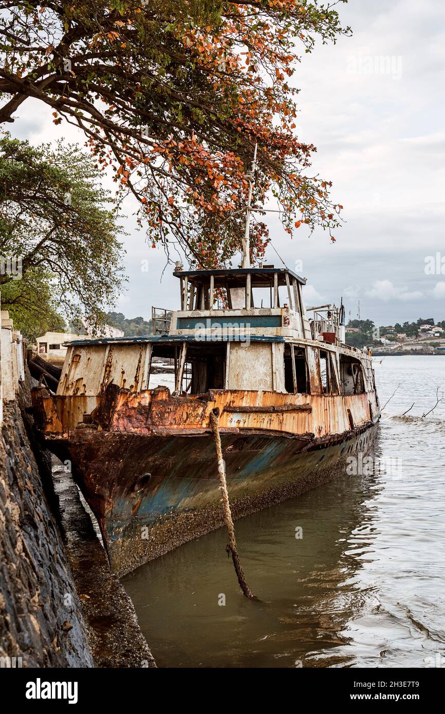 Veraltert verrostet verlassenes Schiff schwimmt in ruhigem Wasser des Golfs von Guinea in der Nähe von Böschung mit Bäumen in São Tomé und Príncipe bei bewölktem Tag bei Tageslicht Stockfoto