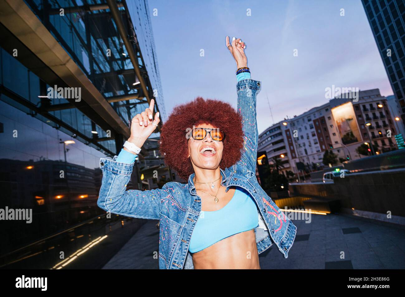 Fröhliche Hündin mit Afro-Frisur in stilvollem Outfit, die abends mit erhobenen Armen auf der Straße mit Wohngebäuden steht Stockfoto