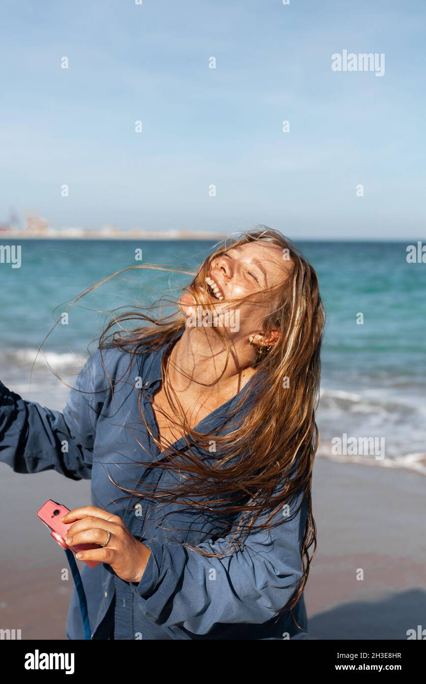 Fröhliche Hündin im nassen Hemd, die am Sandstrand in der Nähe des Meeres steht, während sie an einem sonnigen Tag die Fotokamera hält Stockfoto