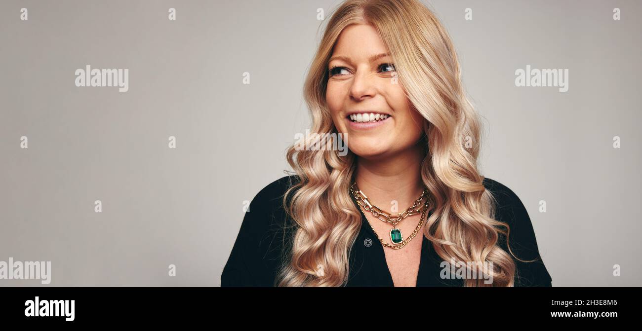 Glückliche Frau, die in einem Studio mit einem Lächeln auf ihrem Gesicht wegschaut. Frau mit mittlerem Erwachsenenalter sieht fröhlich aus, während sie allein vor grauem Hintergrund steht. Stockfoto