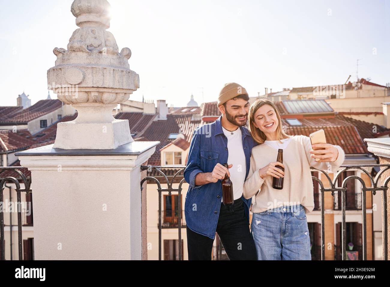 Positive junge Freunde, die zusammen Selfie machen, während sie Bier aus Glasflaschen trinken und sich am Zaun auf dem Balkon lehnen Stockfoto