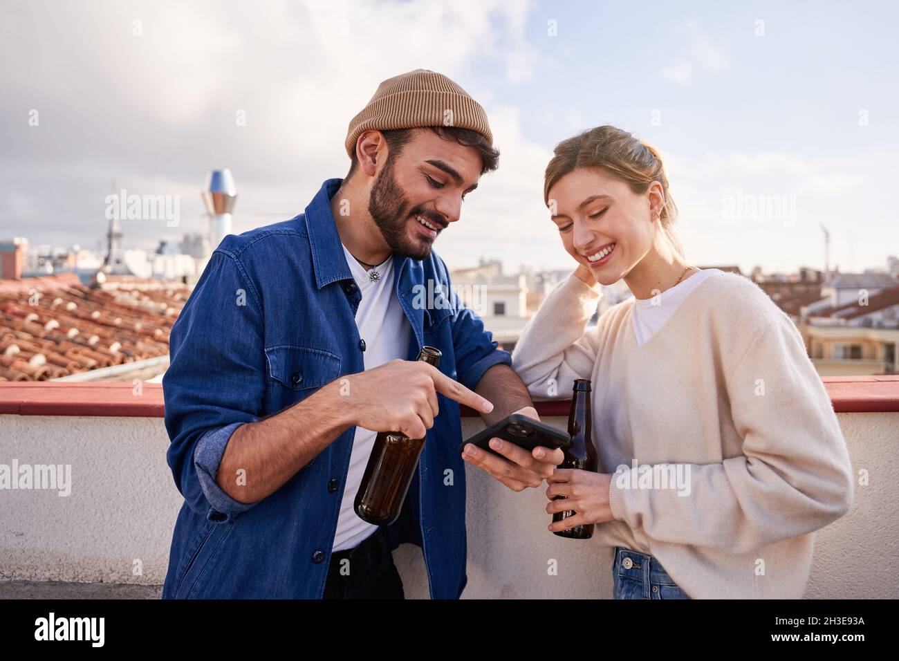 Lächelnder bärtiger Mann mit einer Flasche Bier umarmte die positive Freundin und scrollte am sonnigen Tag auf dem Balkon mit dem Handy Stockfoto