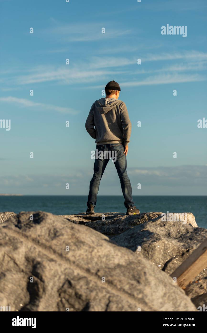 Rückansicht eines Mannes, der auf Felsen steht und auf das Meer blickt, mit einem wolligen Hut und einem Hoodie. Stockfoto