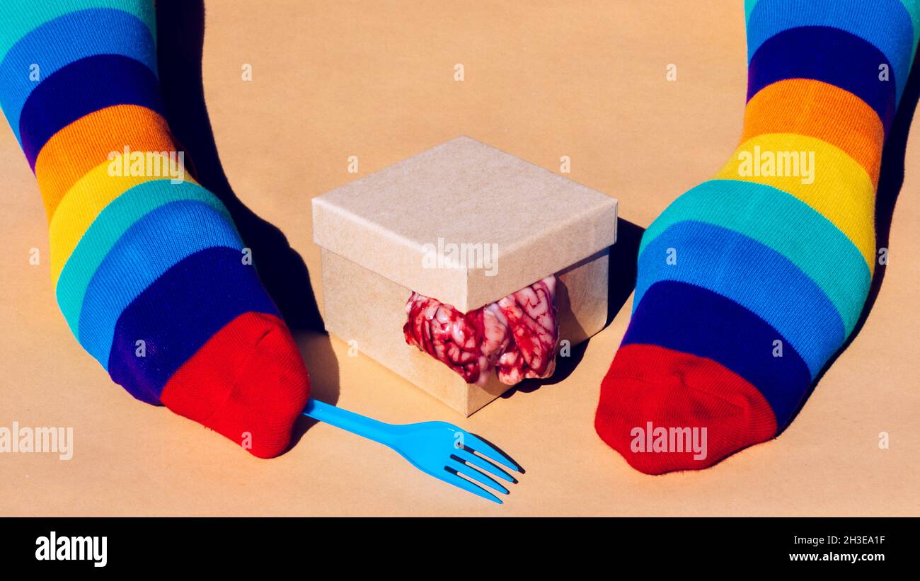 Crop anonyme Person in Regenbogensocken auf Händen mit Gabel in der Nähe von Karton mit rohen Gehirnen auf orangefarbenem Hintergrund im Studio Stockfoto