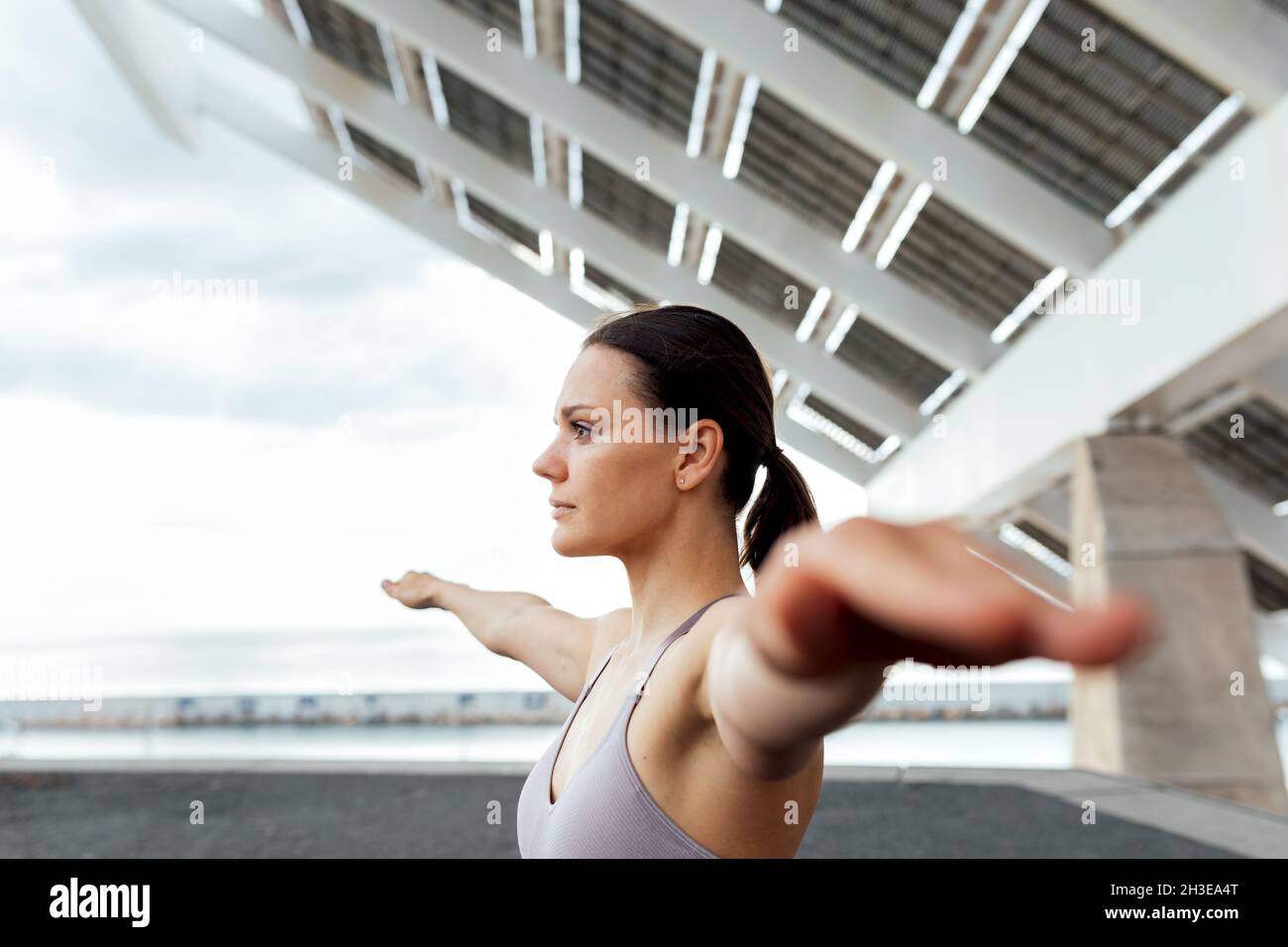 Entschlossenes Weibchen im aktiven Ohr mit ausgebreiteten Armen, die während des Trainings in der Stadt Yoga-Übungen auf der Straße in der Nähe einer modernen Photovoltaik-Anlage machen Stockfoto