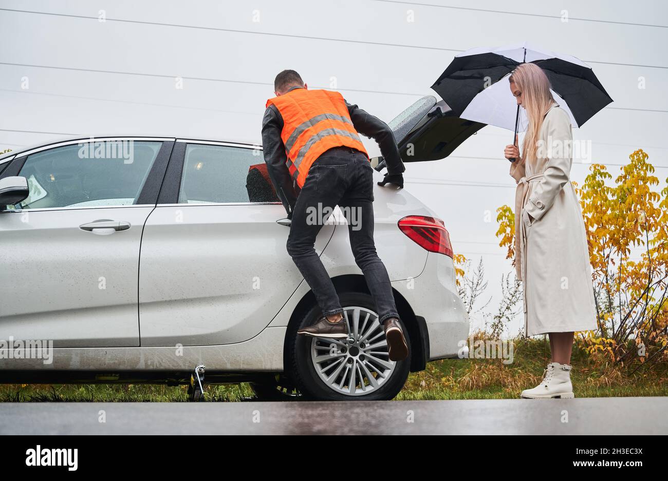 Elegante junge Frau, die neben dem Auto steht und einen Regenschirm hält, während der Automechaniker die Radmuttern abschraubt. Junger Mann in der Weste Reparatur Frau Auto auf der Straße. Konzept des Notdienstes im Straßenverkehr. Stockfoto