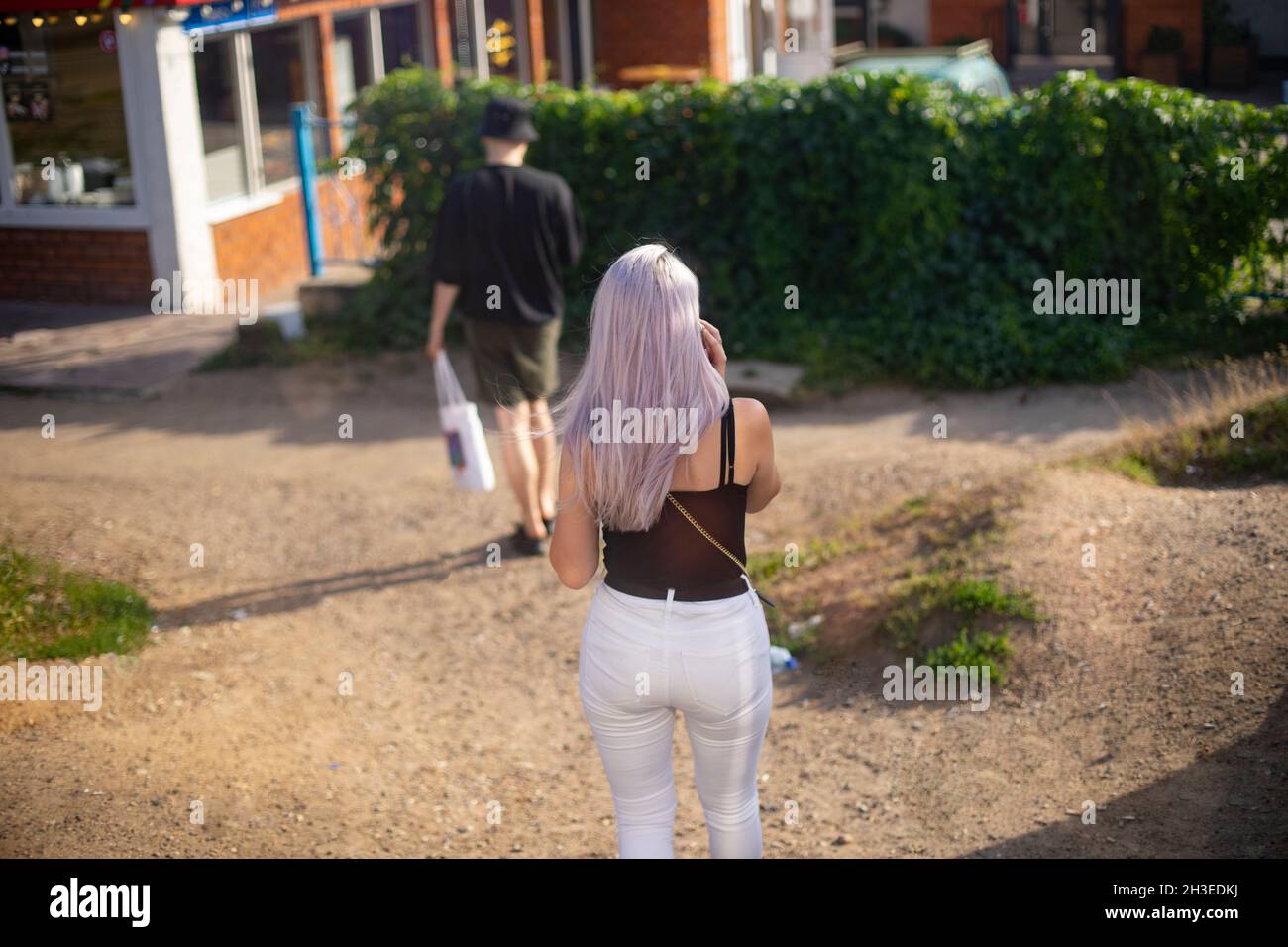 Die Menschen gehen entlang einer sandigen Straße mit eingestreuten Grünflächen im Park. Ein Mädchen mit lila Haaren, in weißen Hosen und einem schwarzen T-Shirt, ein Blick von hinten Stockfoto