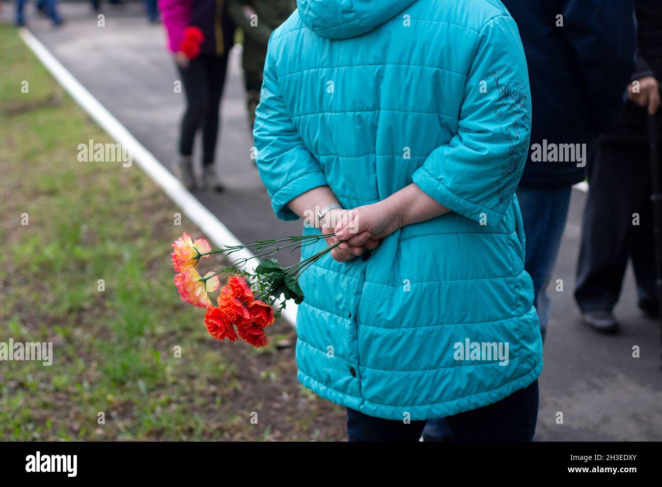 Eine Frau in einer blauen Jacke steht mit Blumen hinter ihrem Rücken. Blick von hinten. Die Straße der Stadt, die Menschenmenge. Tag Des Sieges. Leben In Der Stadt Stockfoto