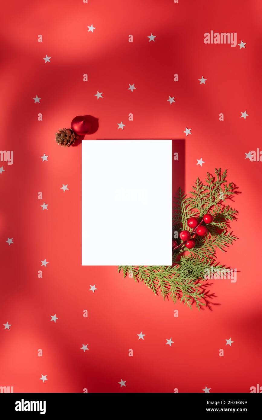 Weihnachtskomposition. Rechteckiges weißes Blatt Papier auf rotem Hintergrund mit Sternen und weichen Schatten, verziert mit Kiefernzweigen, roten Beeren und Ch Stockfoto