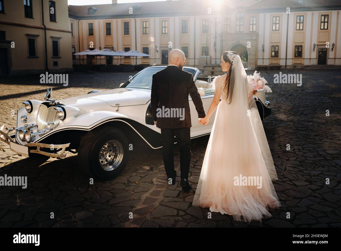 Ein elegantes Brautpaar im Innenhof des Schlosses geht mit den Händen in der Nähe eines Retro-Autos. Stockfoto