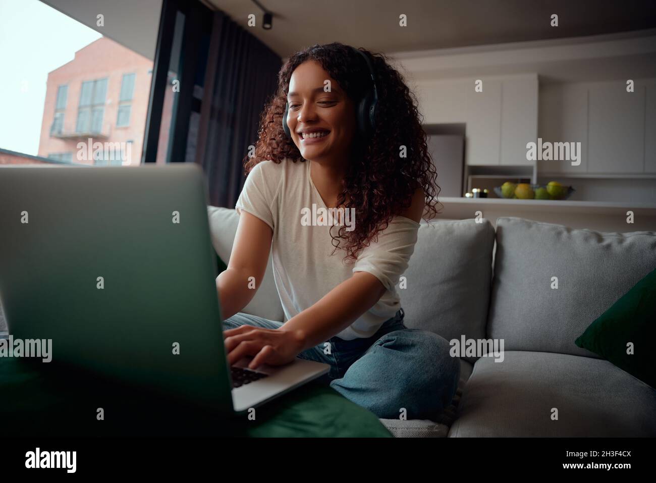 Aufgeregte, multiethnische Frau auf ihrem Laptop, die auf dem Sofa sitzt und Musik hört Stockfoto