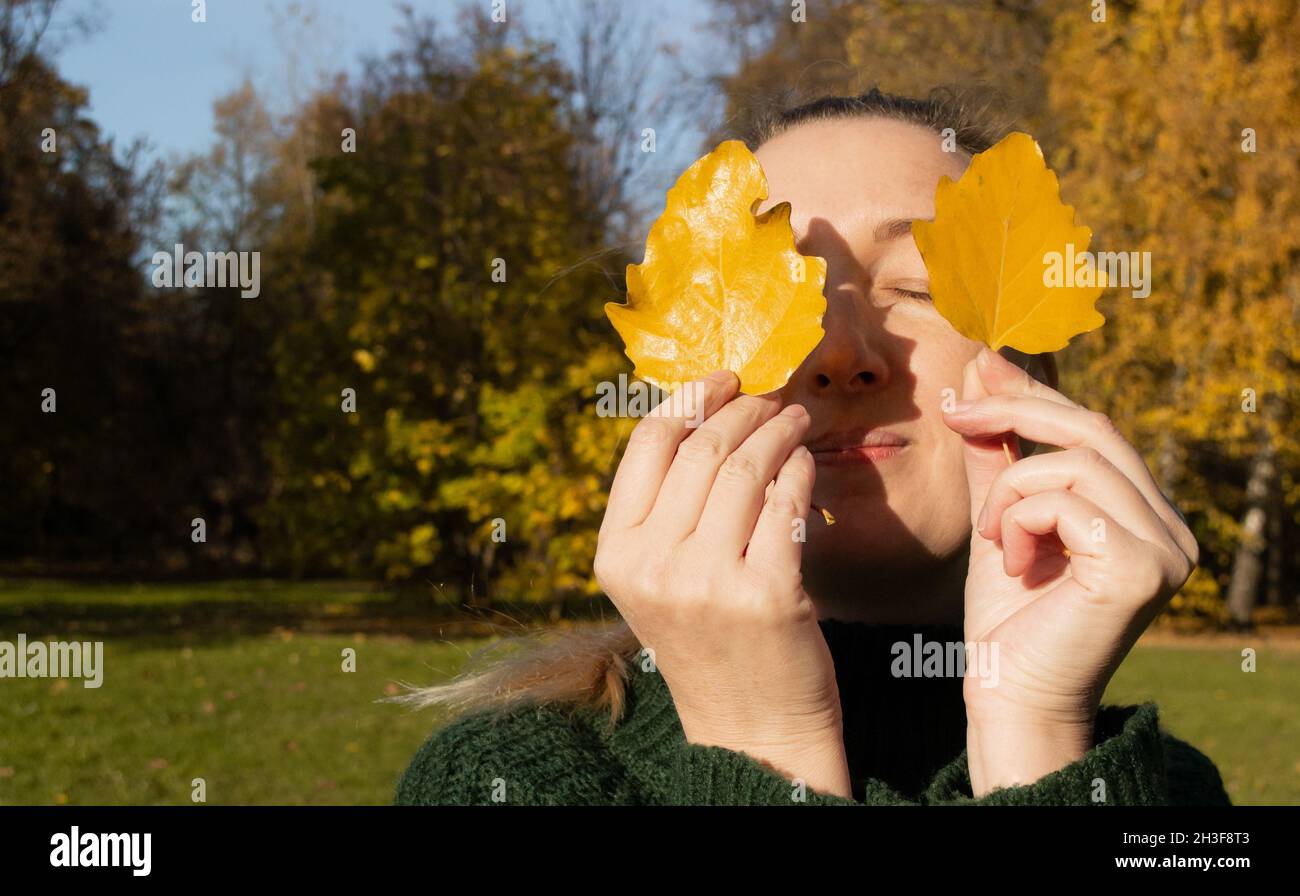 Porträt einer Frau mit geschlossenen Augen hält zwei gelbe Blätter, Hände Nahaufnahme, Hintergrund Herbstbäume. Stockfoto