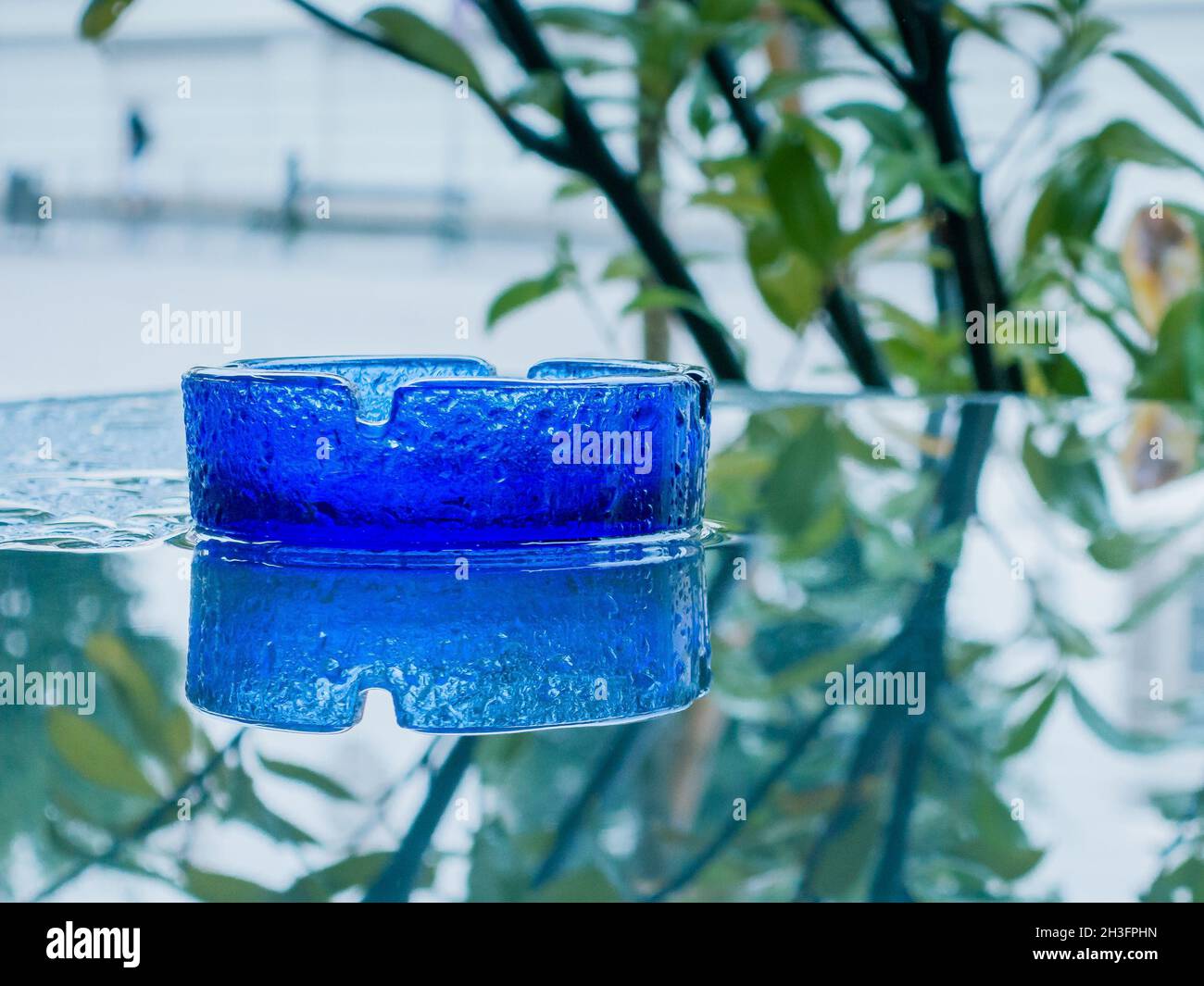 Blaues Glas-Aschenbecher auf nassem Glastisch mit Reflexen Stockfoto