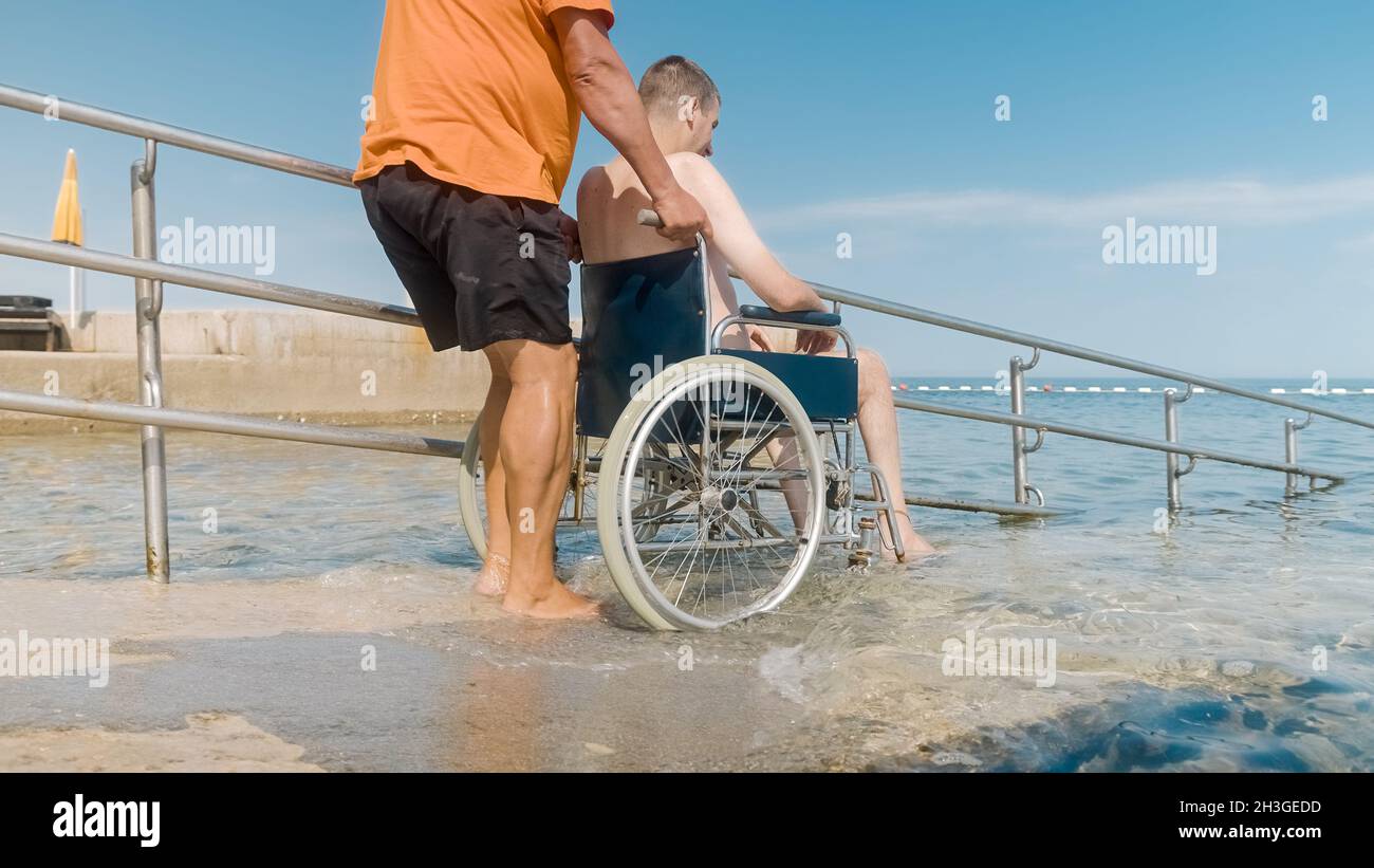 Mann mit Behinderung auf einem Rollstuhl, der zum Schwimmen mit einer Rampe ins Meer transportiert wird. Stockfoto