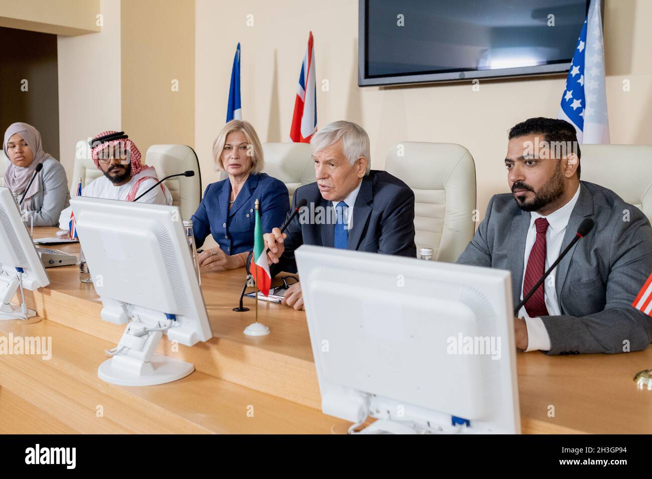 Gruppe von multiethnischen Ländervertretern in Fomalwear, die mit Mikrofonen am Tisch sitzen und auf dem politischen Gipfel globale Probleme diskutieren Stockfoto