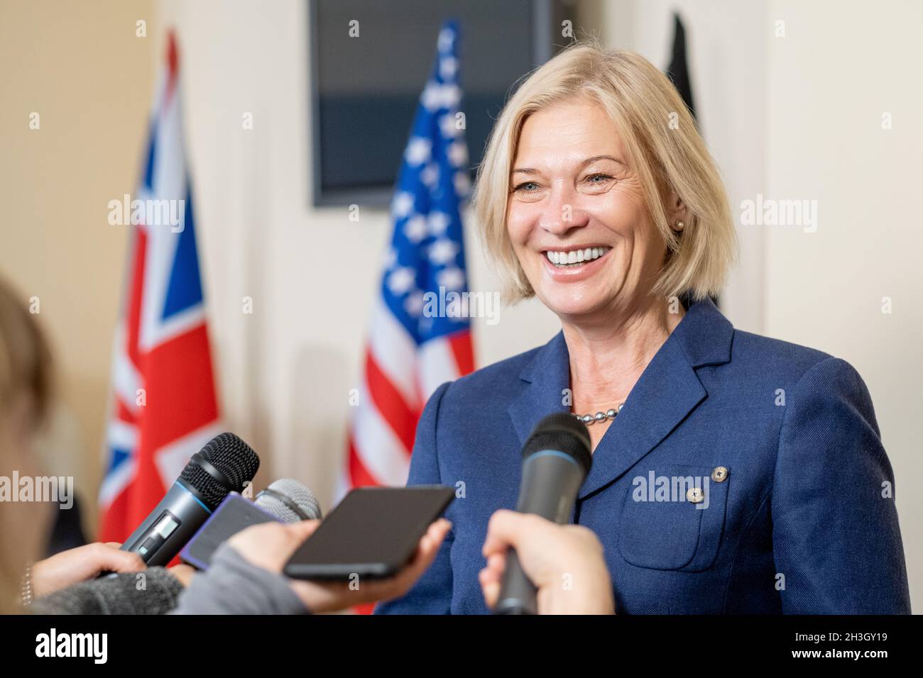 Lächelnde, reife Politikerin mit blonden Haaren, die nach dem Gipfel mit der Presse spricht, während Journalisten Mikrofone erreichen Stockfoto