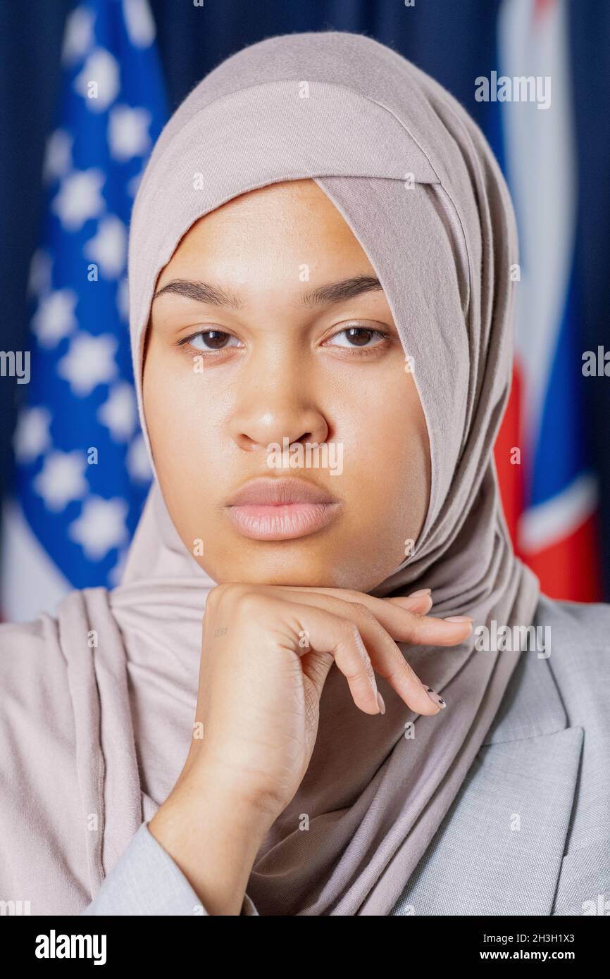 Porträt einer nachdenklichen muslimischen Politikerin in Hijab, die sich mit dem Kopf an der Hand gegen Nationalflaggen lehnt Stockfoto
