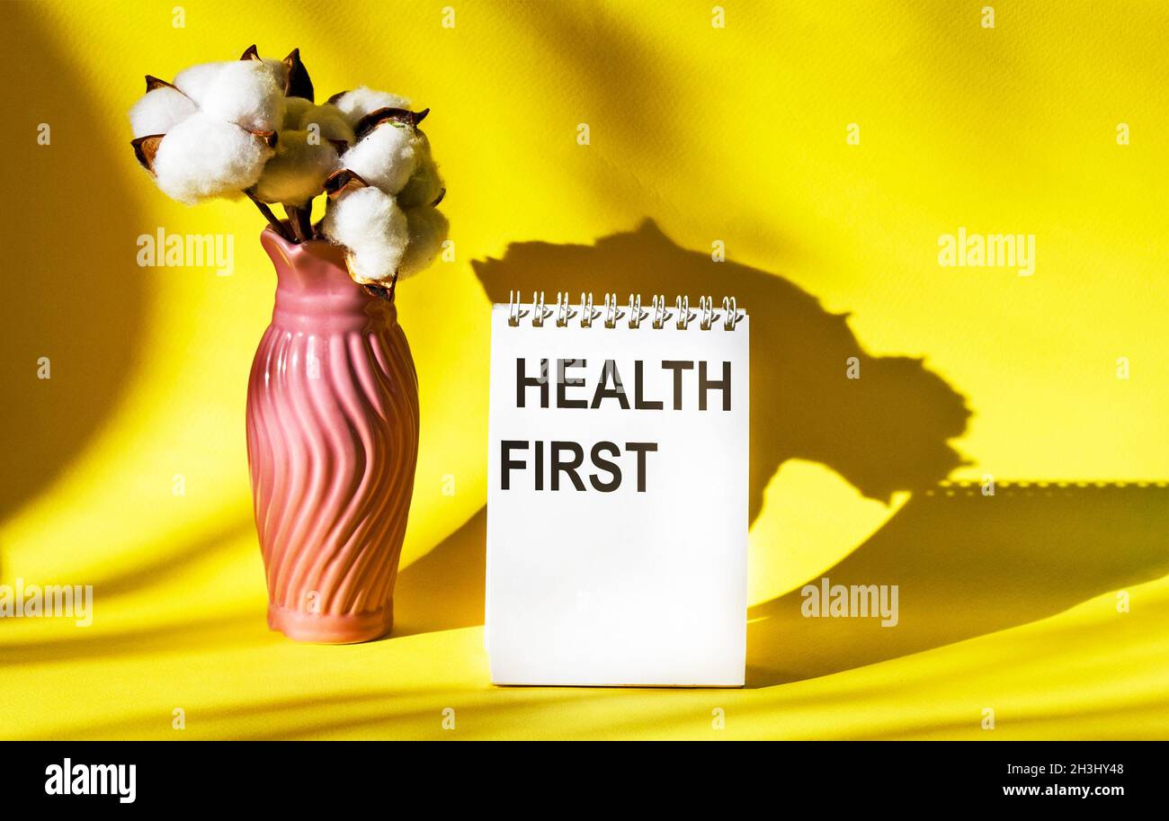 Das Wort GESUNDHEIT STEHT ZUERST für einen Notizblock auf einem gelben Hintergrund mit einem Bund Baumwolle. Medizinisches Konzept. Stockfoto