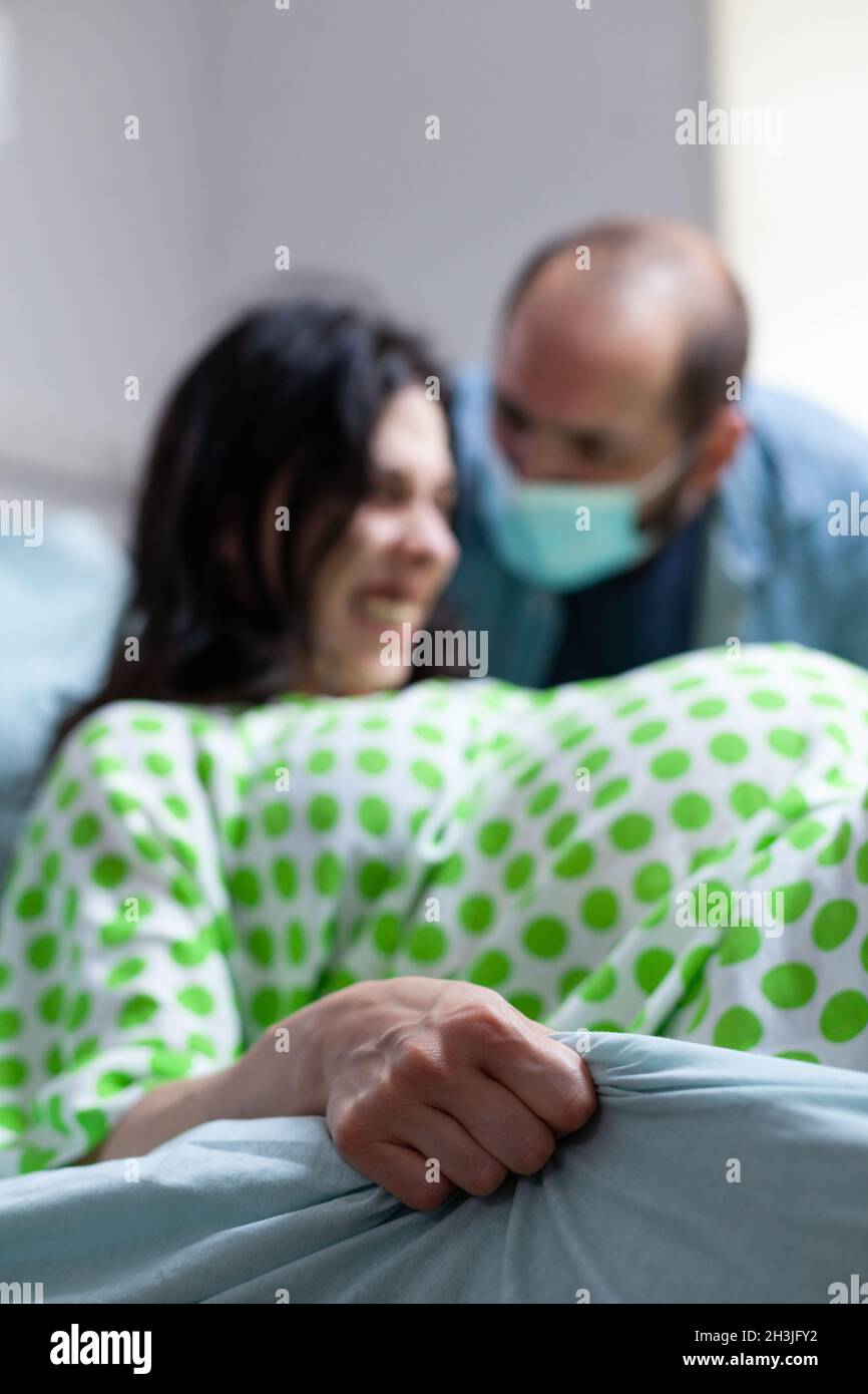 Nahaufnahme einer Frau, die das Bettlaken mit der Hand hält, während sie gebiert und schmerzhafte Kontraktionen auf der Krankenstation hat. Person in der Kindergeburten Druck und Ehemann bei der Mutterschaft zu helfen Stockfoto