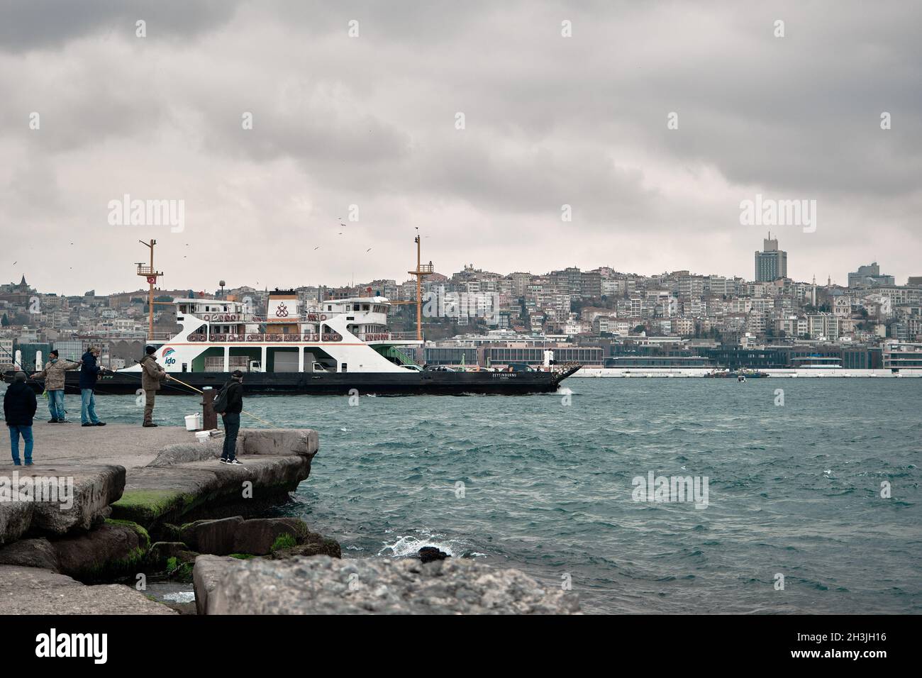 Fußgängerfähre, die in bosporus istanbul in der Nähe des sarayburnu-Viertels vorbeifährt, mit vielen Menschen, die bei bewölktem Wetter an der Küste spazieren und angeln Stockfoto
