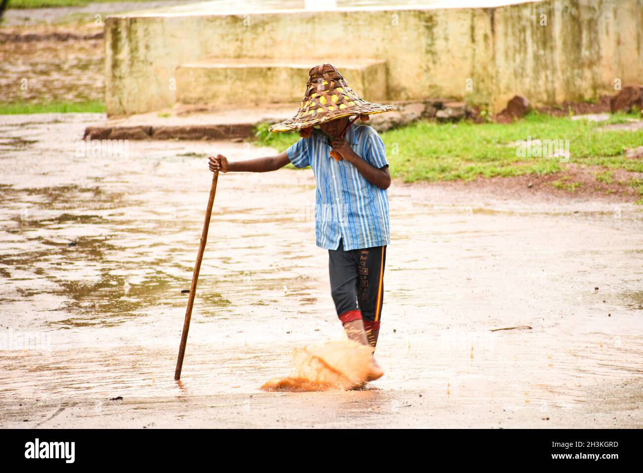 SURAJPUR, INDIEN - 01. Apr 2019: Ein kleiner Dorfjunge in einem Strohhut mitten auf der Straße, der während des Regens mit Wasser spielt Stockfoto