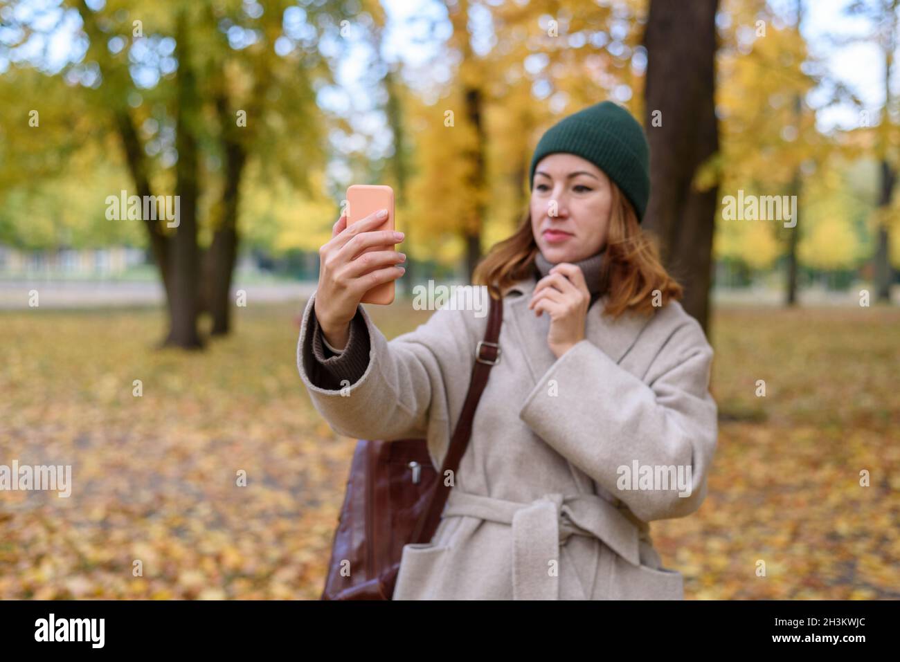 Frau, die ihre Haare oder ihr Aussehen fixiert und durch die Frontkamera wie einen Spiegel im Herbstpark auf den Bildschirm schaut Stockfoto