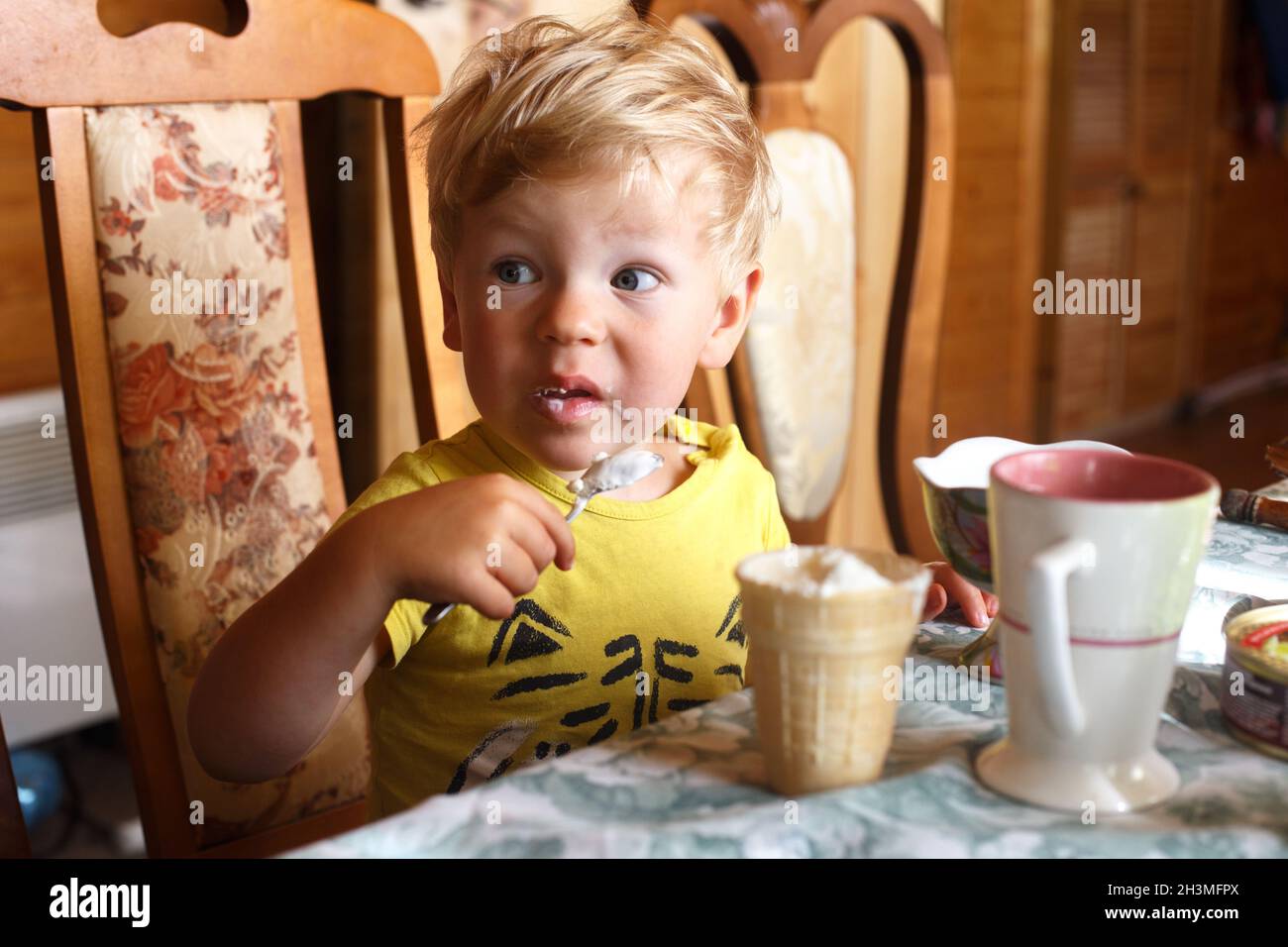 Ein blonder Junge sitzt am Tisch und isst selbst mit einem Löffel Eis Stockfoto