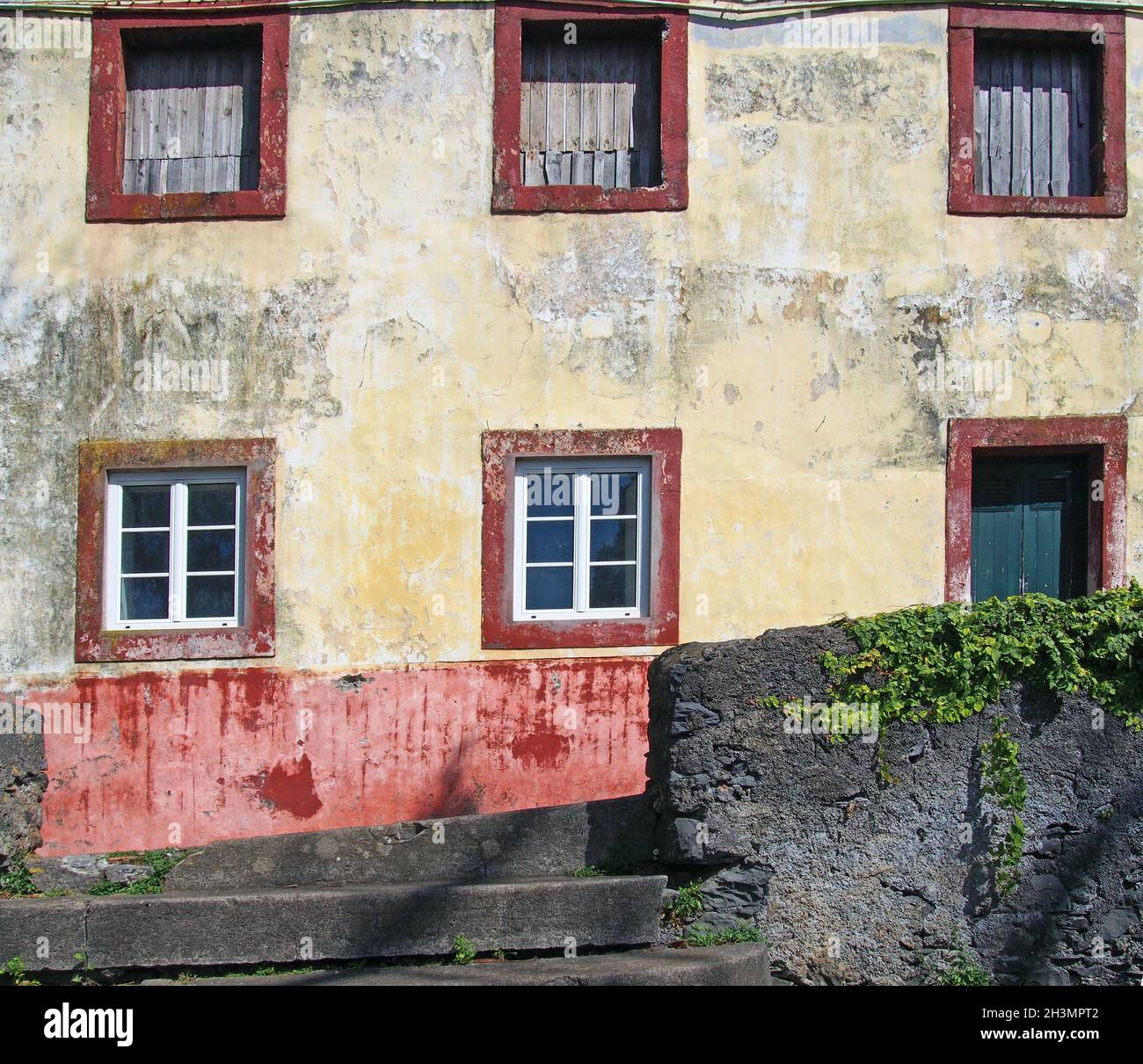 Fassade eines typischen alten spanischen Hauses in verblassten Gelb- und Rottönen, umgeben von einer grauen Wand mit Pflanzen in Funchal, T Stockfoto