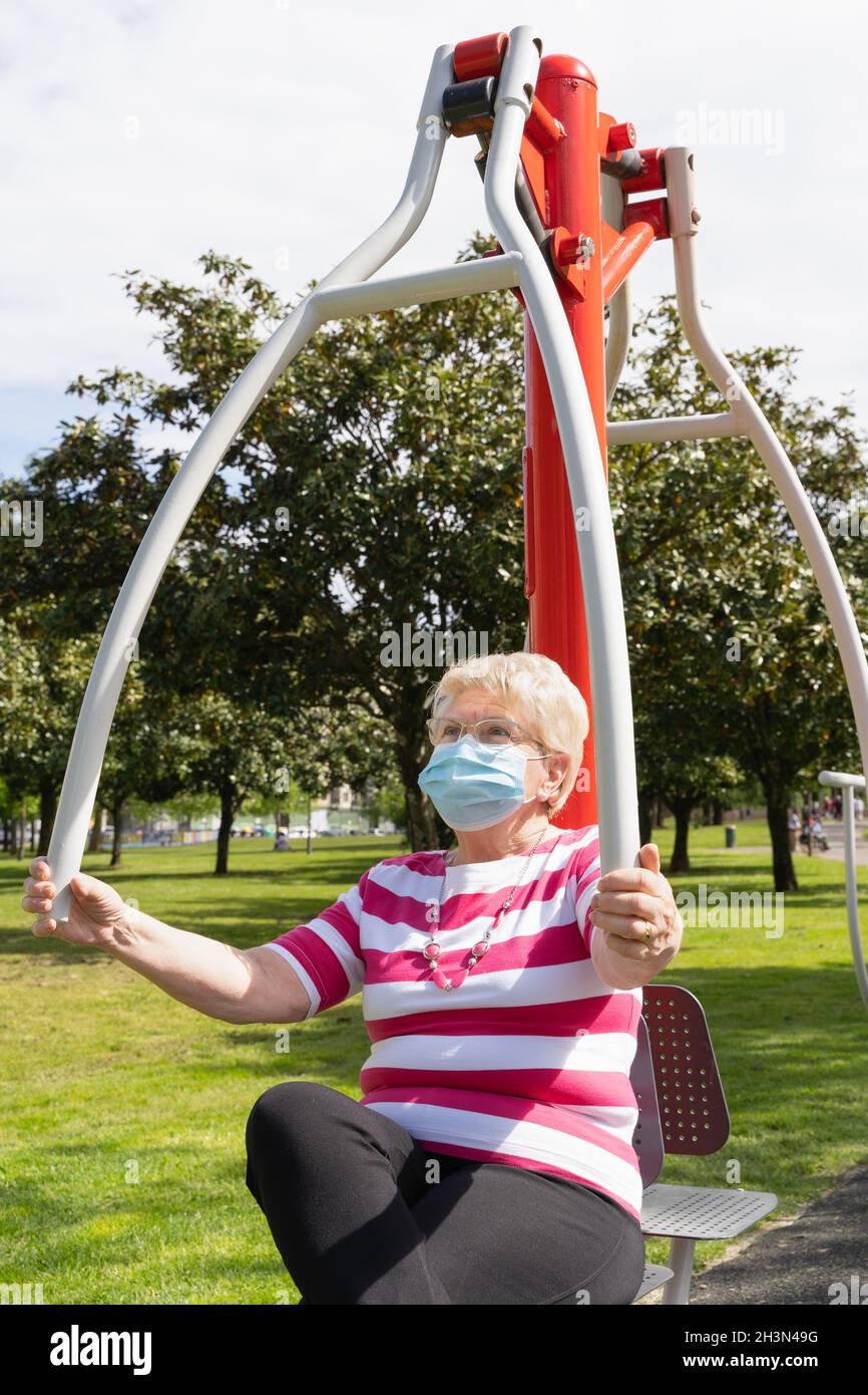 Ältere blonde Dame, die mit Gesichtsmaske und Brille körperliche Aktivitäten am Park-Trainingsgerät macht. Ältere Frau trainiert den Oberkörper, um sich fit zu halten Stockfoto