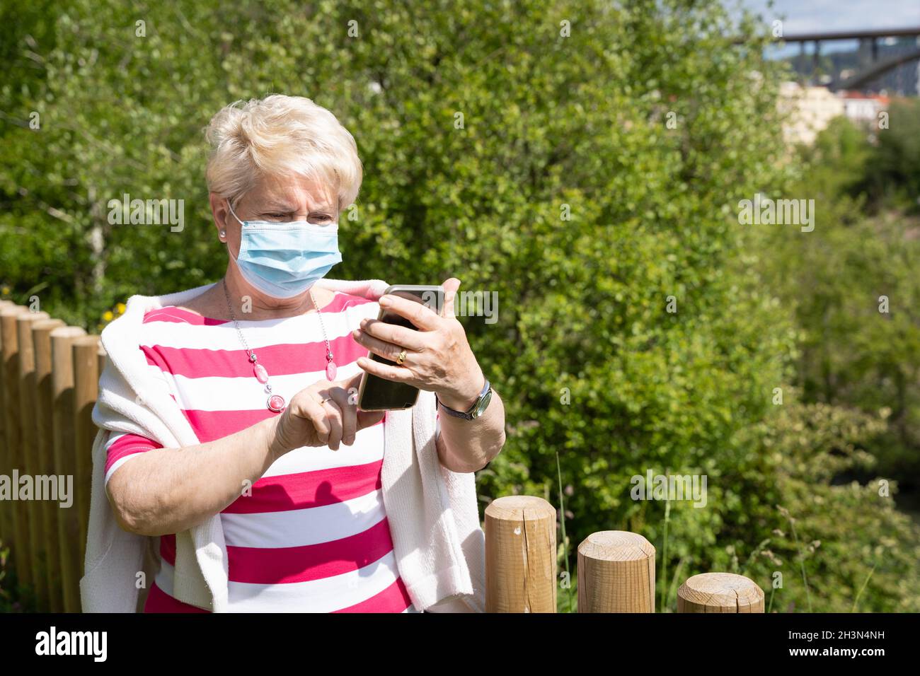 Ältere Frau mit Gesichtsmaske, die am sonnigen Tag den Handybildschirm im Park berührt. Ältere Dame, die mit dem Mobiltelefon auf der natürlichen Landschaft steht Stockfoto