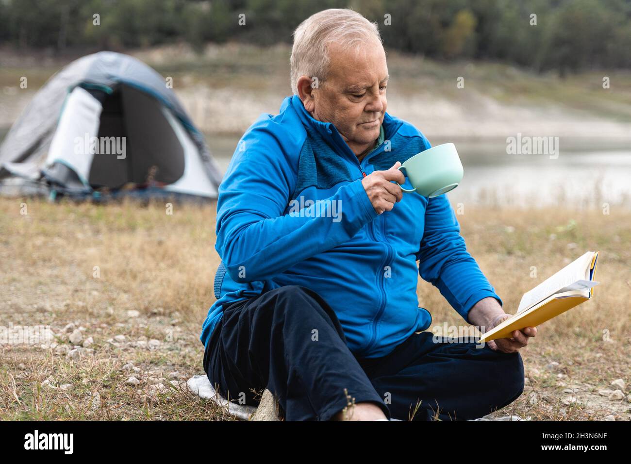 Älterer Mann, der mit Zelt zeltet, während er Buch liest und Kaffee mit See im Hintergrund trinkt - echte ältere Menschen Stockfoto