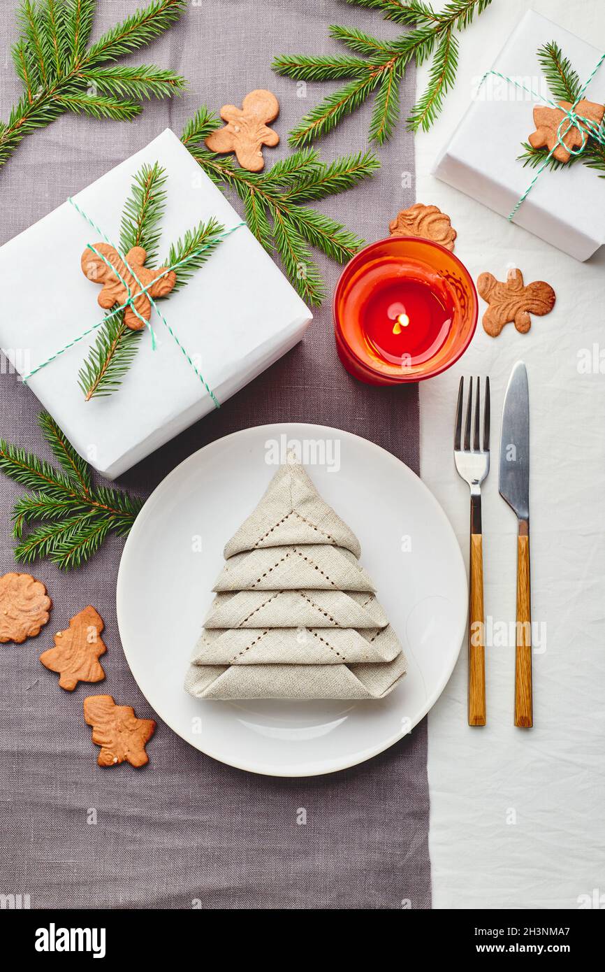 Serviette in Form eines Weihnachtsbaums auf einem Teller auf weißer Tischdecke mit Geschenken und Dekorationen mit Tannenzweigen und Lebkuchen Stockfoto