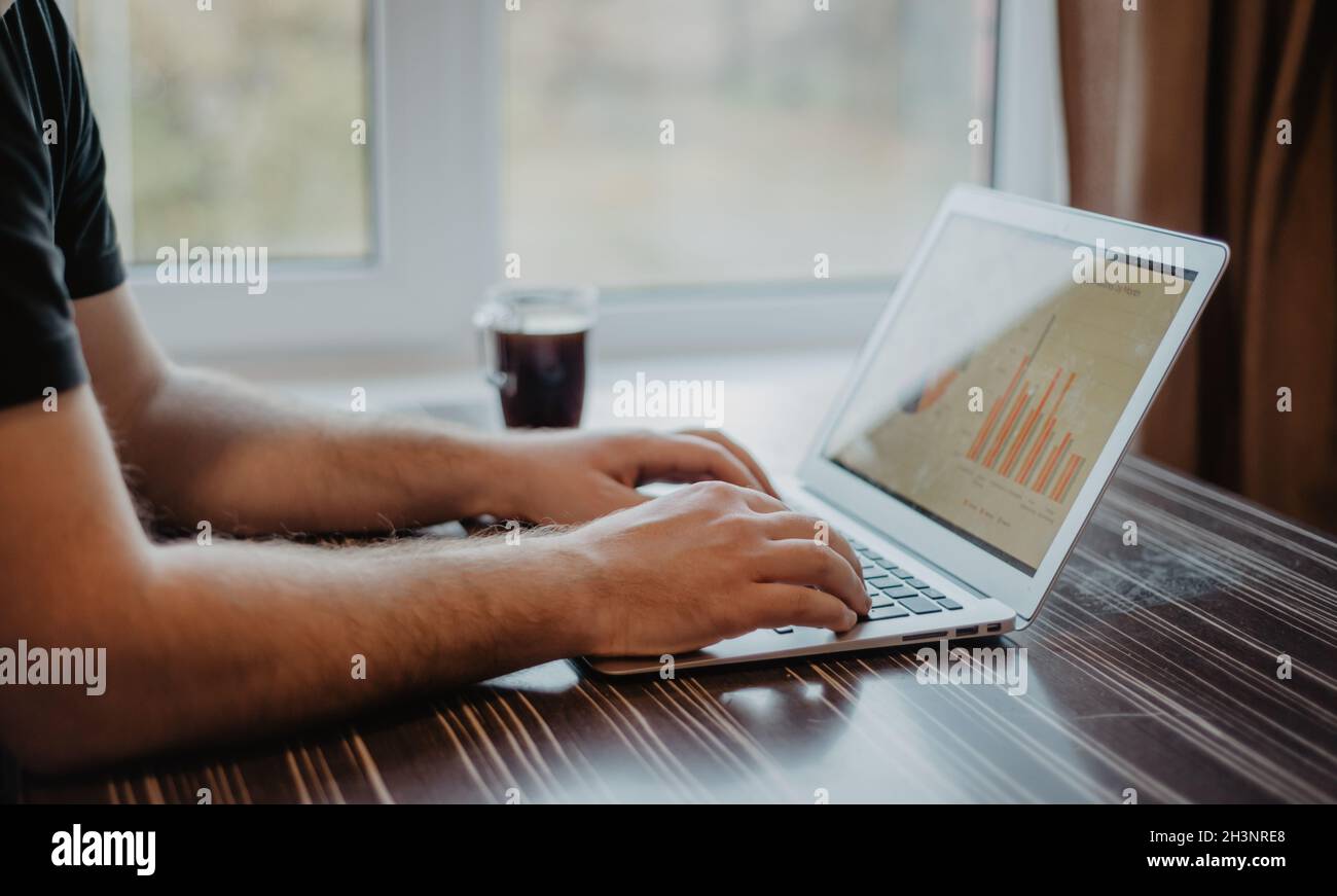 Nahaufnahme eines Mannes, der auf einem Laptop tippt und mit einem Finanzdiagramm auf einem unordentlichen Bürotisch aus Holz arbeitet. Stockfoto