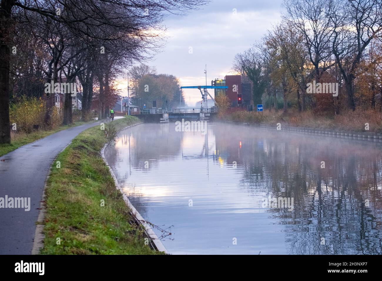 Flusskanal, der sich mit Grasbänken und wilden Blumen und Bäumen in einer malerischen Landschaft bei einem nebligen Herbstmorgen-Sonnenaufgang biegt Stockfoto