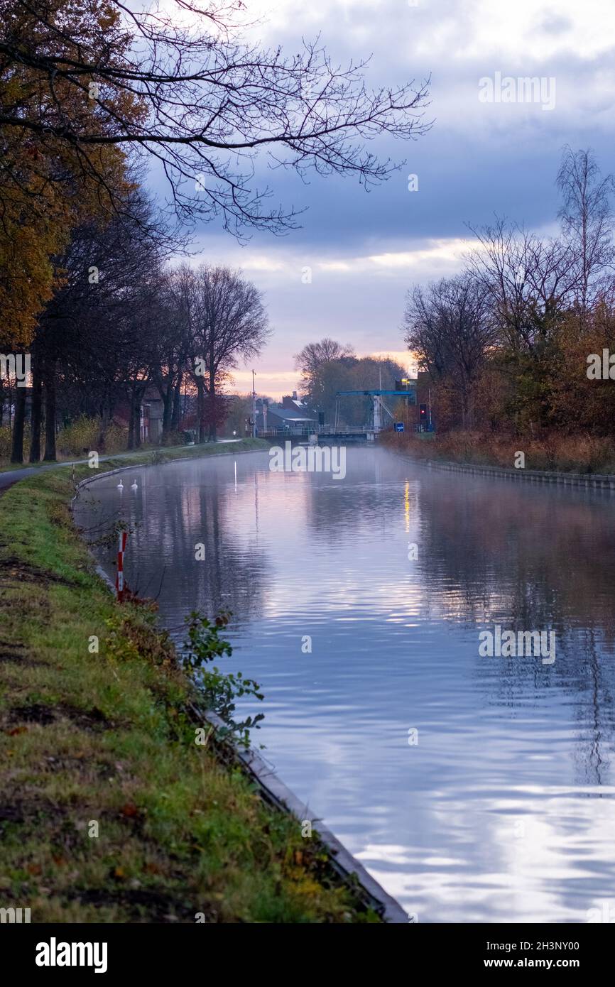 Flusskanal, der sich mit Grasbänken und wilden Blumen und Bäumen in einer malerischen Landschaft bei einem nebligen Herbstmorgen-Sonnenaufgang biegt Stockfoto