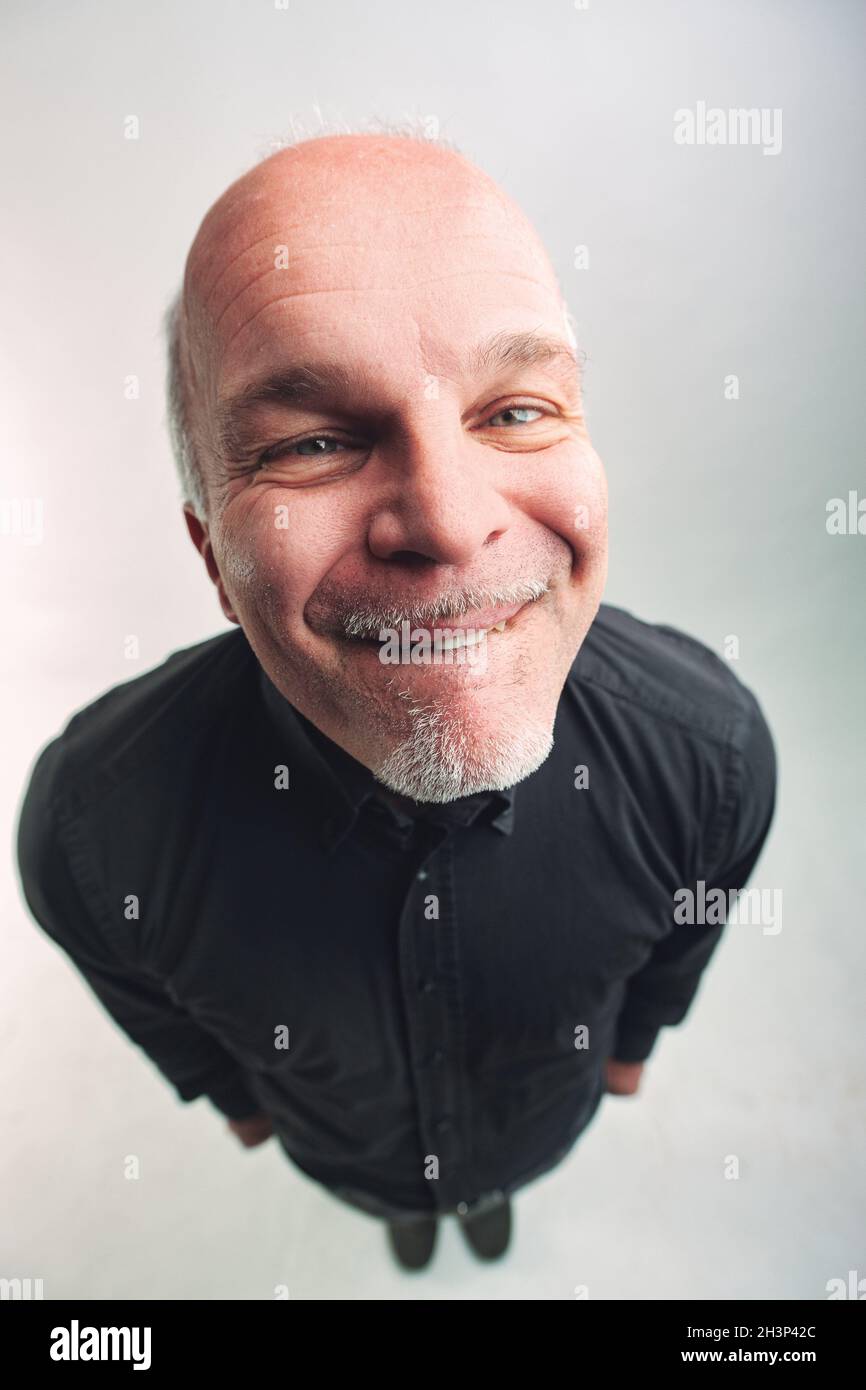 Draufsicht auf einen schrulligen älteren Mann, der sich auf die Lippe beißt, während er mit amüsed lächelnden Augen über einem weißen Studiohintergrund auf die Kamera blickt Stockfoto