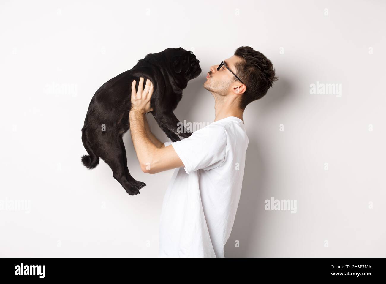 Profil von schönen jungen Mann küssen kleinen niedlichen Hund Gesicht. Hipster Kerl liebt seinen Mops, stehen über weißem Hintergrund Stockfoto