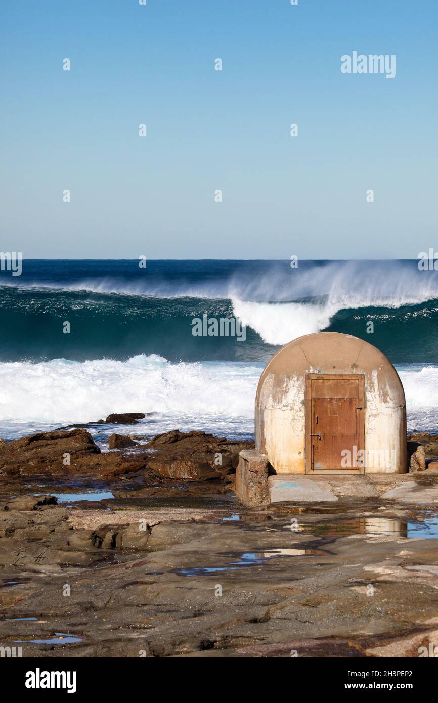 Große Brandung, die hinter dem Pumphouse am Cowrie Hole - Newcastle Australia bricht. Dieses Pumphouse ist ein Wahrzeichen der Region und liefert Wasser in die Umgebung Stockfoto