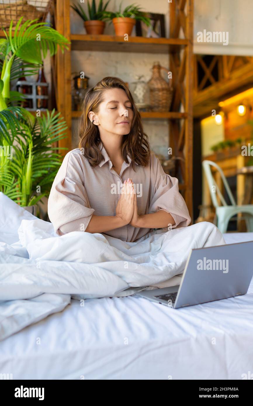Konzept der Wiederherstellung von Kraft und geistiger Energie durch Yoga-Praxis - junge Erwachsene Frau meditiert im Bett vor dem Laptop-Monitor. Vertikales Foto Stockfoto
