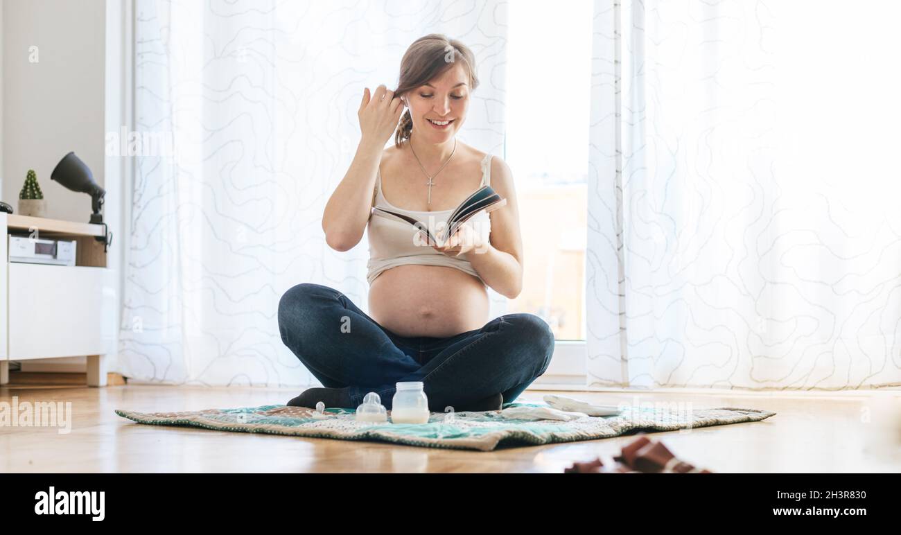 Lesen über Schwangerschaft: Eine glückliche, schwanger Mutter sitzt auf dem Boden und liest ein Buch Stockfoto