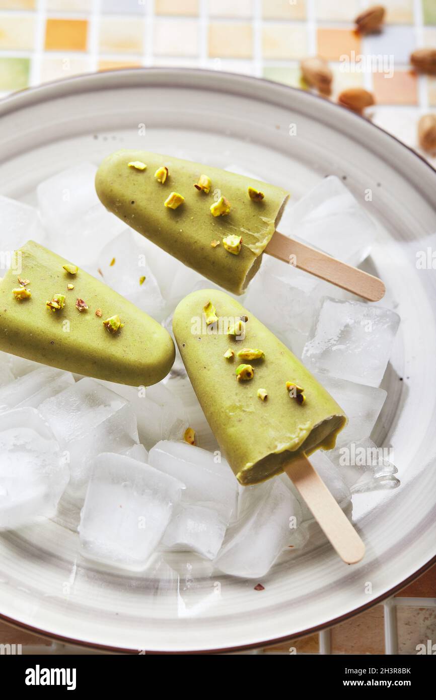 Gefrorener hausgemachter Pistazien-Popsicle in einer Schüssel mit Eis auf einem Tisch mit Mosaikfliesen. Erfrischender Fruchteis, gefrorener grüner Saft auf Stick. Oben vi Stockfoto