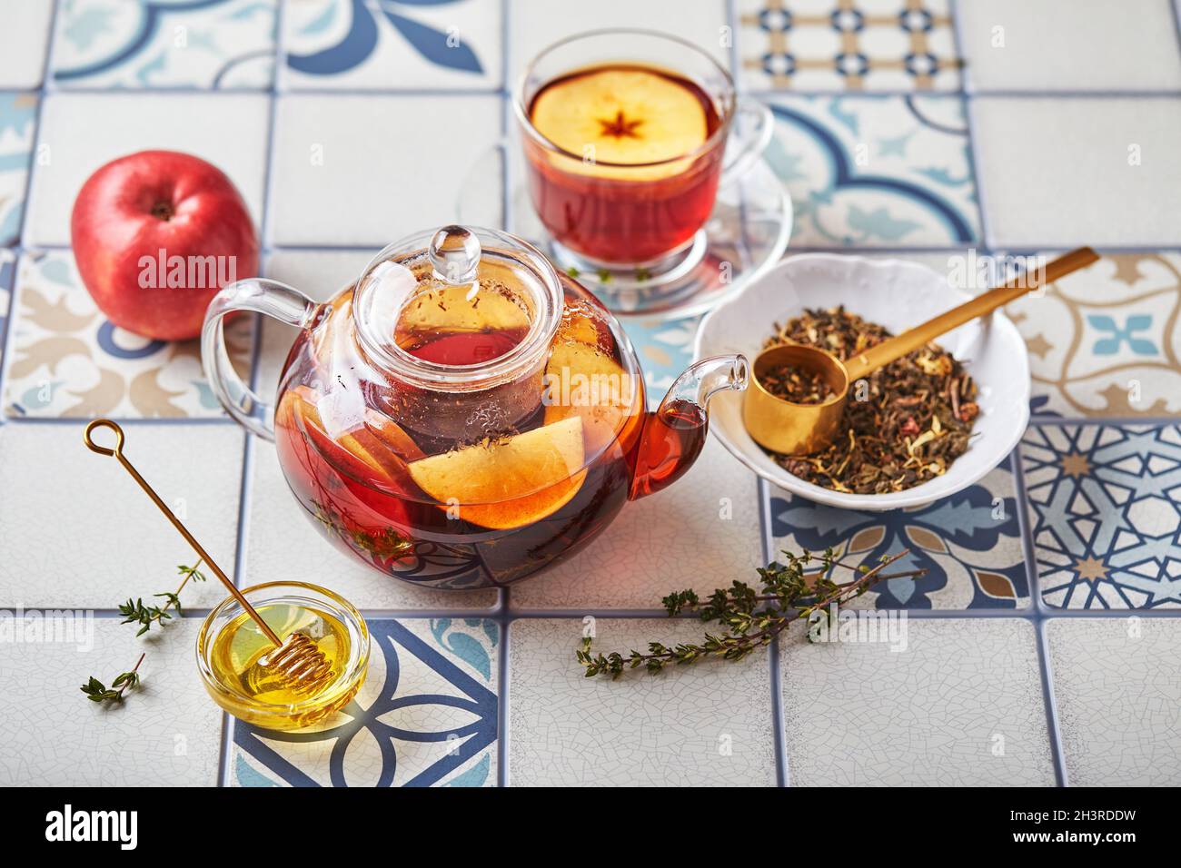 Früchtetee mit Äpfeln und Thymian in Glas Teekane und Tasse auf Tisch aus farbigen Fliesen Stockfoto