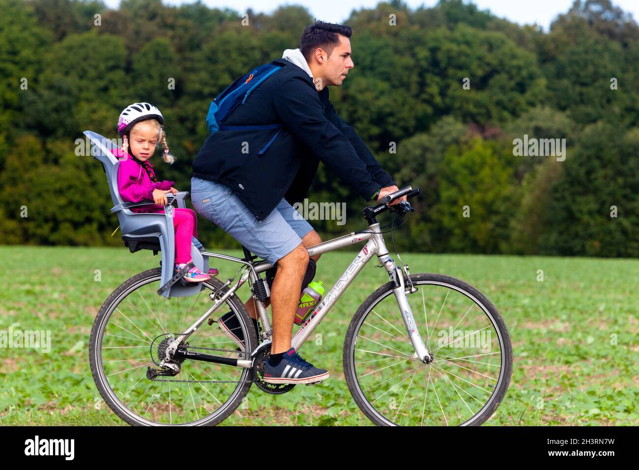 Ein kleines Mädchen im Kindersitz, ein Mann auf dem Fahrrad, ein Kind auf dem Fahrradsitz, ein Kleinkindhelm Stockfoto