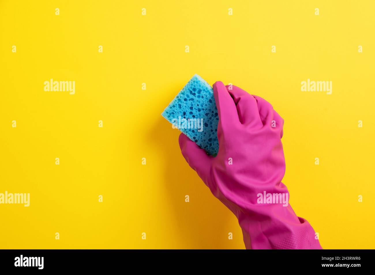 Eine Hand in einem rosa Gummihandschuh hält einen blauen Waschschwamm auf gelbem Hintergrund Stockfoto
