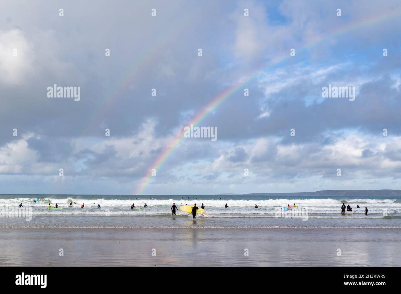 Surfer am Strand in Newquay Cornwall wunderschöner Sommertag mit einem echten doppelten Regenbogen über dem Meereshorizont. Viel Surfen und Surfbrett Stockfoto