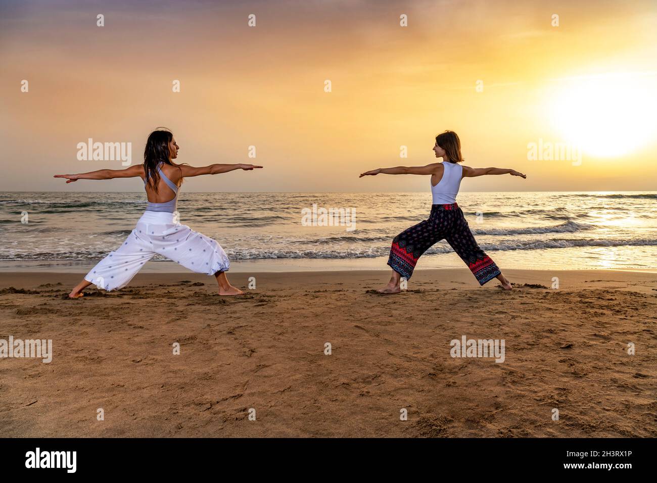 Zwei junge Frauen, die den Kriegeryogagang machen, posieren am Strand bei Sonnenuntergang. Gefühl von Energie und Stabilität in einer Spätsommeratmosphäre Stockfoto