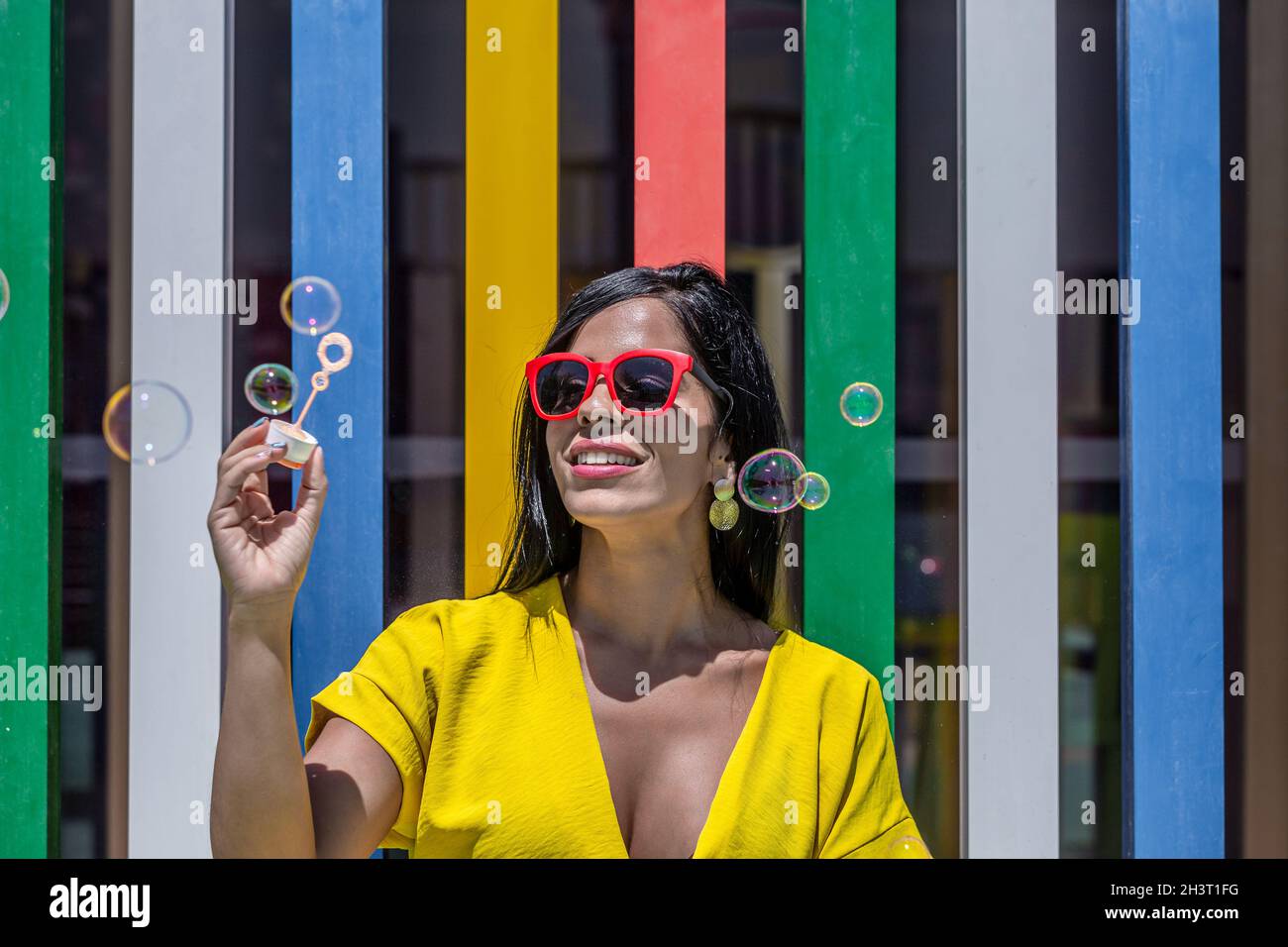 Vorderansicht einer Latina-Frau mit Bubbles Zauberstab in einem gelben Kleid und Sonnenbrille vor einem Hintergrund von bunten Bars an einem sonnigen Tag. Stockfoto